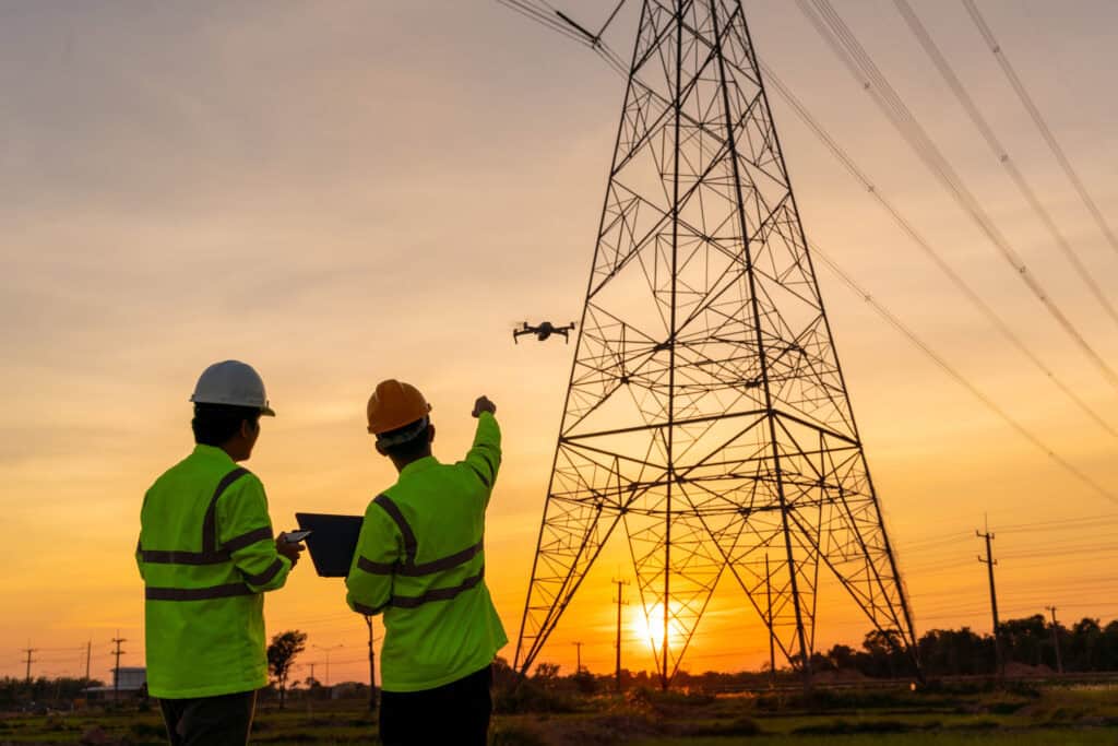 Two engineers in helmets and hi-vis jackets pointing at a drone near an electric transmission tower at sunset